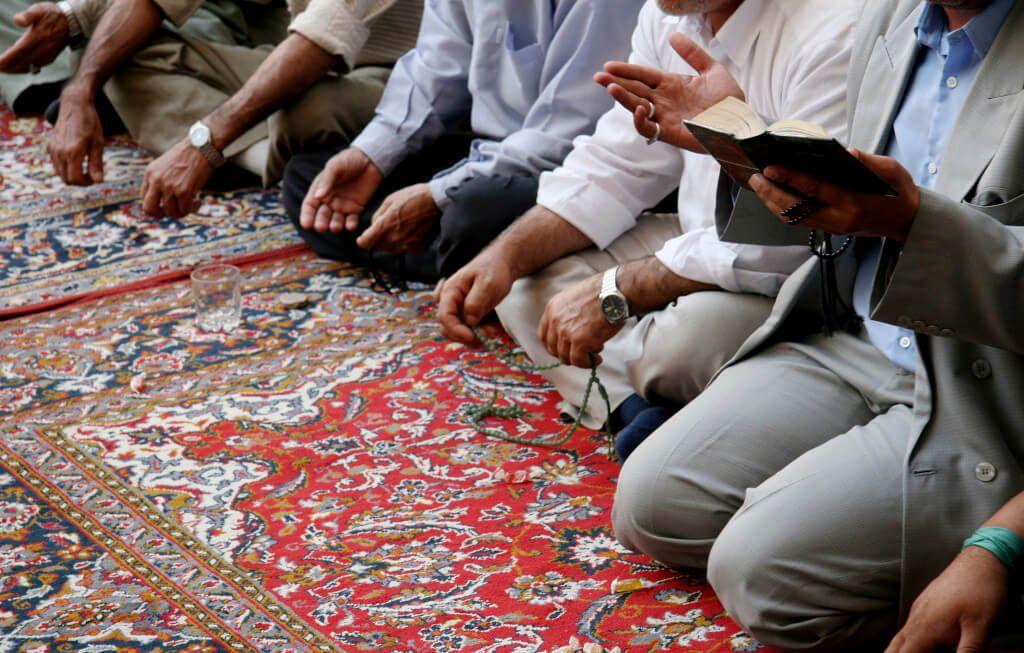 Several people sitting on a colorful carpet, holding prayer beads and reading from a book.